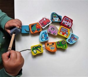 Obrázek zobrazuje dítě navlékající dřevěné kostky, které jsou vymalované jako dopravní prostředky.